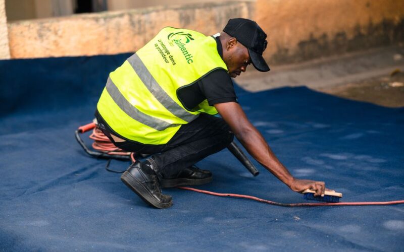 A worker dressed in a safety vest cleans a blue carpet outdoors with a brush.