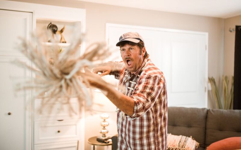 A surprised man with a checkered shirt holding a mop indoors, expressing intense emotion.