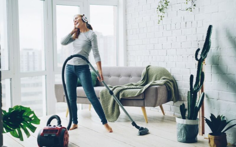 A cheerful woman listens to music on headphones while vacuuming in a bright, modern living room.