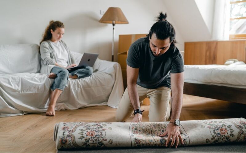 Fit ethnic male in wristwatch laying carpet on parquet while barefoot woman browsing internet on portable computer sitting on cozy sofa in bedroom