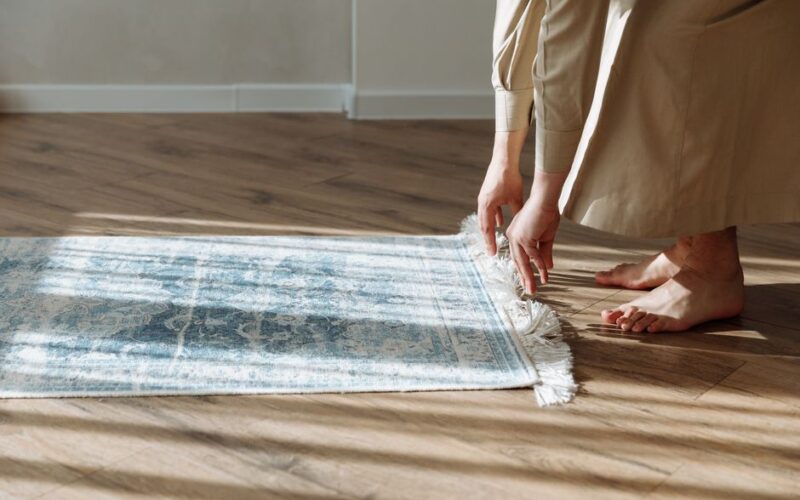 Person adjusting a prayer rug on a sunlit wooden floor, highlighting peaceful homemaking.