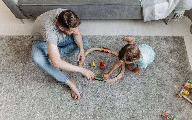 Father and young son playing together with a toy train set on a carpeted floor at home.