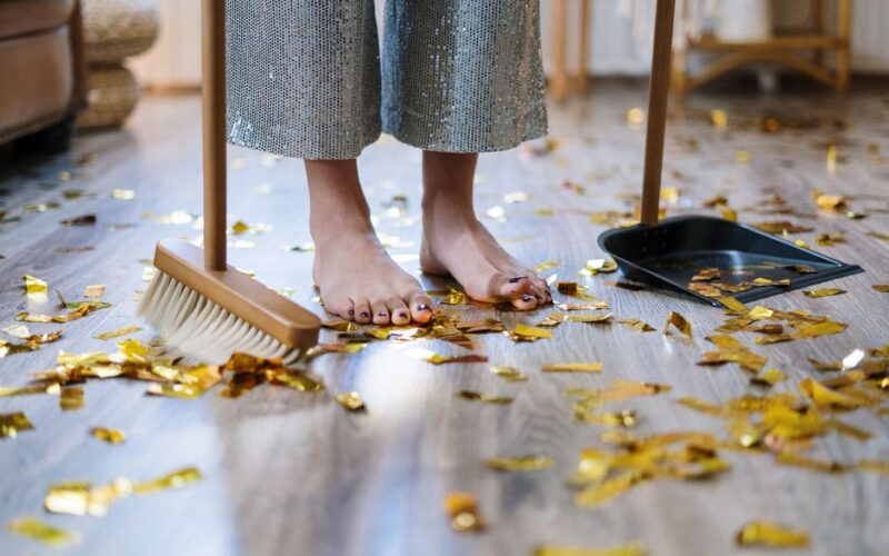 Barefoot woman sweeping gold confetti with broom and dustpan indoors.
