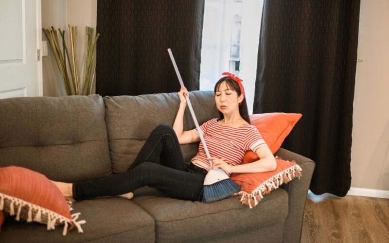 Woman lounging on a sofa holding a broom, in a cozy indoor setting.