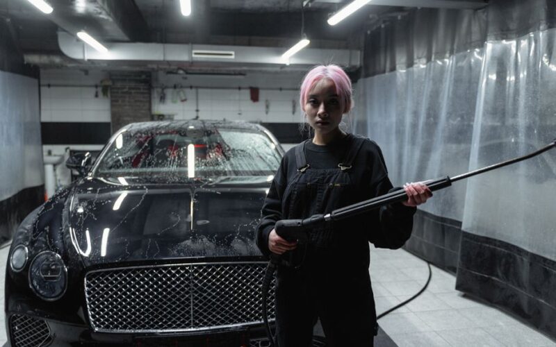 Person using a pressure washer to clean a luxury black car in an indoor garage.
