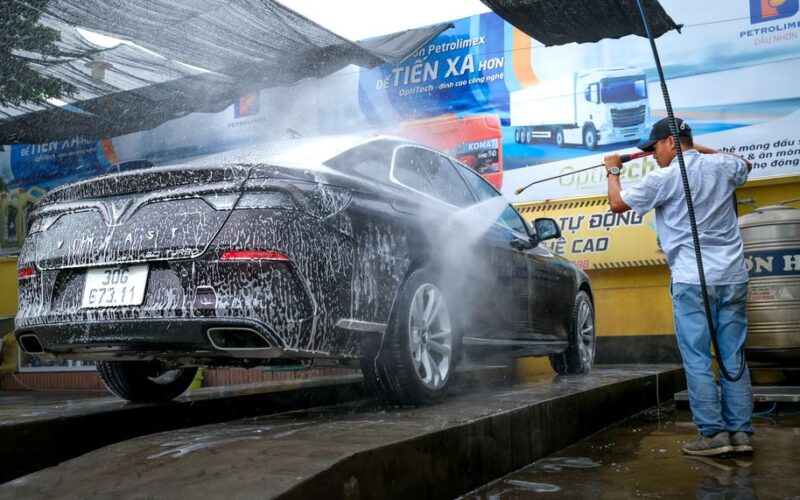 A black car is being cleaned with a power spray by a man at a car wash station outdoors.