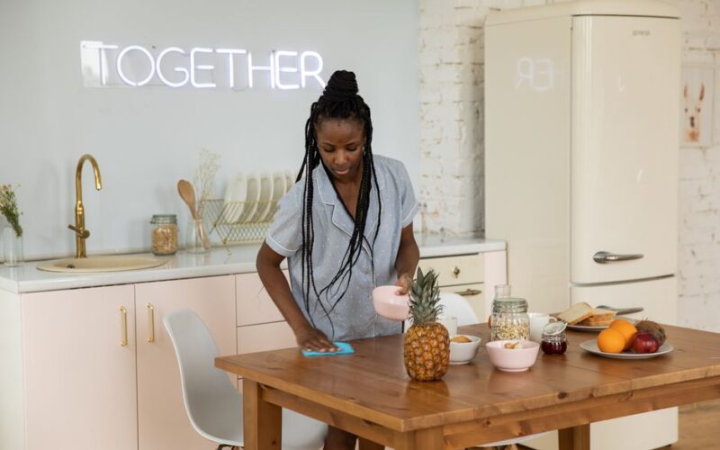 African American woman tidying up a kitchen table with fresh fruits and breakfast items, showcasing a clean dining area.