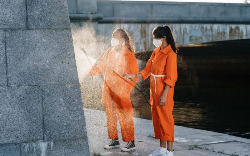 Two people in orange jumpsuits performing outdoor cleaning while wearing face masks.