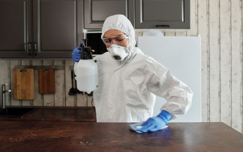 A person in protective clothing disinfects a kitchen counter indoors, focusing on safety and cleanliness.